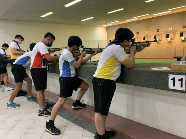 Several young athletes in colorful jerseys aim rifles at targets in an indoor shooting range.